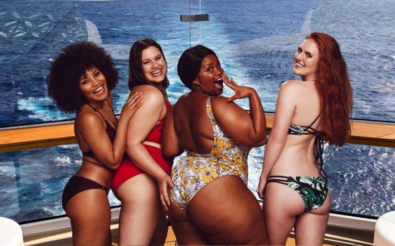 Four women in swimsuits pose playfully on a cruise ship, with the ocean visible through large windows behind them. They are smiling and embracing, showcasing body positivity and diverse beauty.