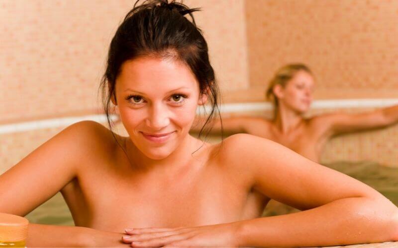 Two women are relaxing in a hot tub, with one smiling at the camera and the other reclining in the background. The setting appears serene and spa-like, with warm tones and a calm ambiance.