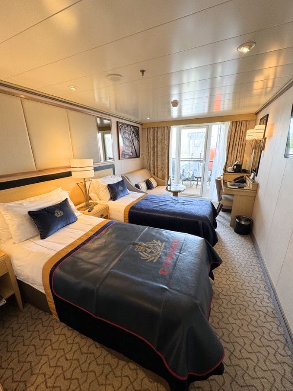 A Cunard cruise ship stateroom with an obstructed view balcony, featuring two neatly made twin beds with navy blue bedding, a desk with a lamp, and a small seating area near the balcony door.