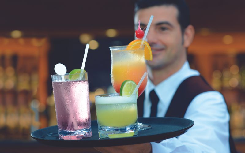 A waiter holding a tray with three colorful cocktails, each garnished with fruits and served with straws. The background is blurred, highlighting the vibrant drinks.