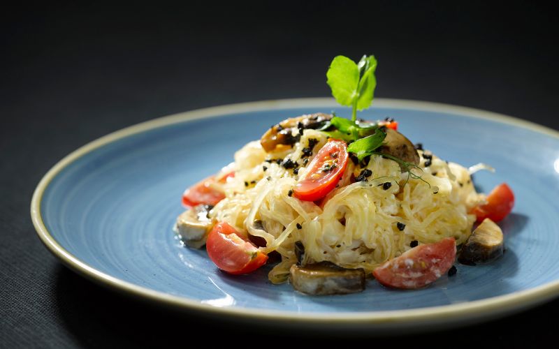 A plate of gourmet pasta garnished with cherry tomatoes, mushrooms, black sesame seeds, and a sprig of green herbs. The dish is presented on a blue plate against a dark background.