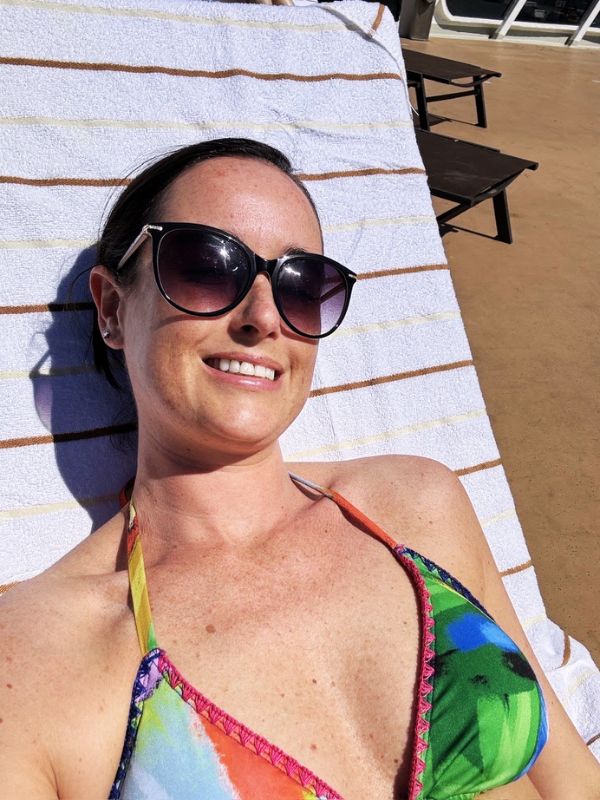 Cruise Mummy wearing sunglasses and a colorful bikini relaxes on a lounge chair covered with a striped towel on the deck of the Cunard Queen Mary cruise ship. She is enjoying the sunny weather, lying on a spacious deck area with additional lounge chairs in the background.
