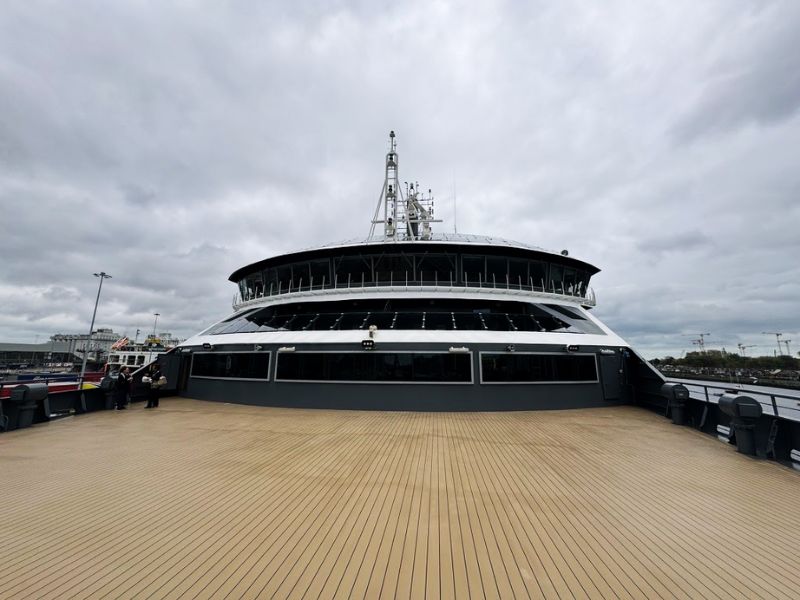 Expansive outdoor deck area on the Seabourn Venture, showcasing a wide, open space with a sleek wooden floor that sets a luxurious tone. The deck surrounds the ship's navigation bridge, which is visible in the center with its large, circular structure filled with windows for panoramic viewing. The deck provides ample room for guests to stroll, relax, and take in views of the port under a vast, cloudy sky, highlighting the ship’s robust and stylish design. This area is ideal for enjoying the fresh air and scenery during the voyage.