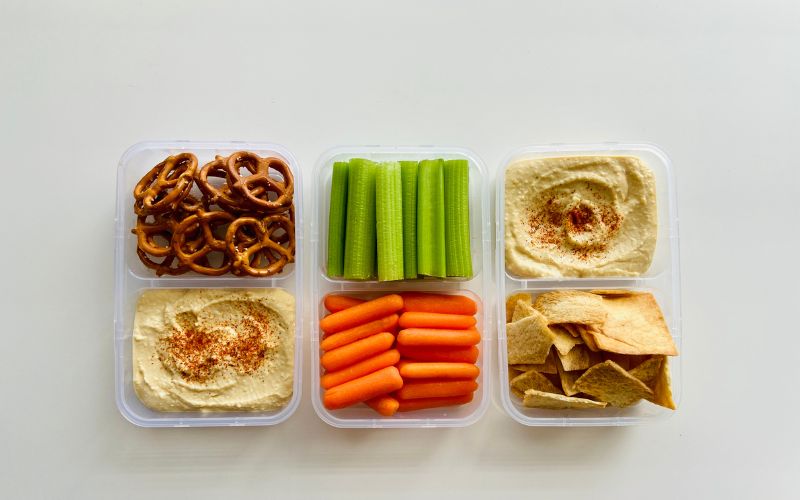 A plastic container filled with hummus, pretzels, carrots, and celery, perfect for a healthy snack on the go.