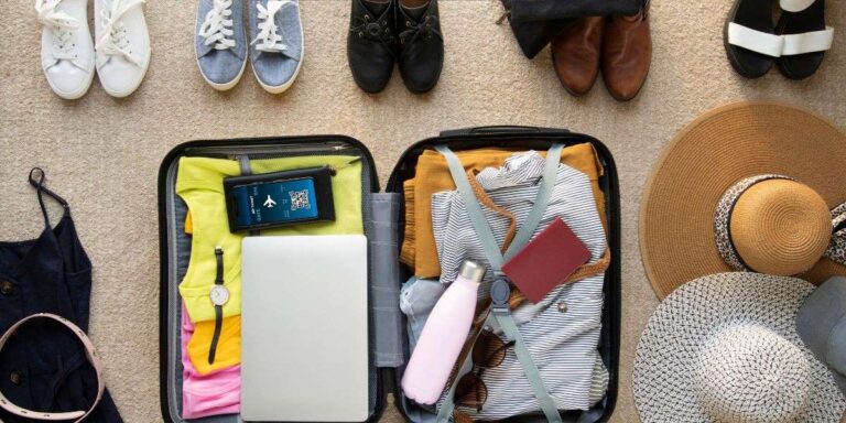 An open suitcase packed for a cruise trip shows neatly organized clothing, a passport, a water bottle, a watch, and a laptop. Surrounding the suitcase are various shoes, a sun hat, and other travel essentials, all laid out on the floor in preparation for the journey.