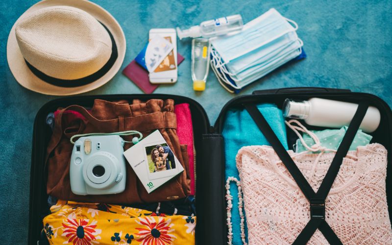 Open suitcase packed with travel essentials: colorful clothing, instant camera with photo, sun hat, face masks, hand sanitizer, and travel documents, symbolizing readiness for a safe and enjoyable journey.