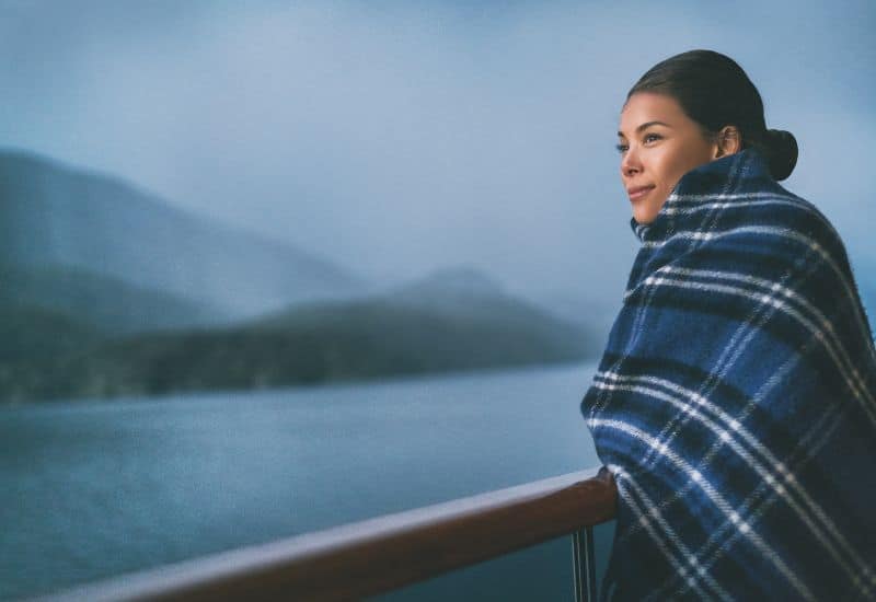 A passenger wrapped in a plaid blanket stands on the deck of an Alaskan cruise ship, gazing out at the serene, misty landscape.