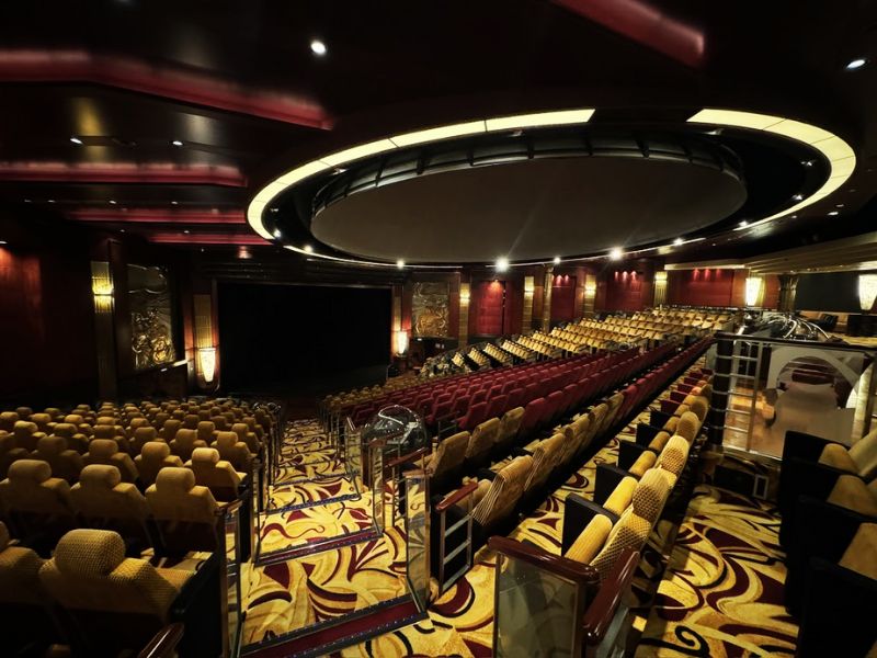 The interior of the planetarium on the Cunard Queen Mary cruise ship features rows of plush, patterned seats facing a central stage and screen. The room is adorned with ornate decorations and has a large circular ceiling design, creating a luxurious and immersive atmosphere.