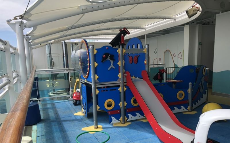 Outdoor play area on a cruise ship, featuring a nautical-themed play structure with slides, climbing elements, and interactive features. The area is covered with a canopy for shade, providing a fun and safe environment for young children to play and explore while at sea.