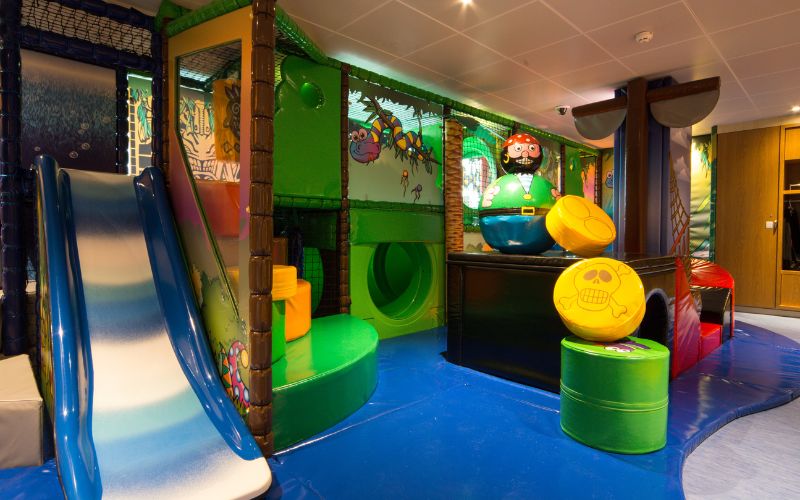 Colorful indoor play area on a cruise ship, featuring a slide, climbing structures, and pirate-themed decorations. The room is designed for young children with various interactive play elements and soft, cushioned surfaces, providing a safe and fun environment for active play.