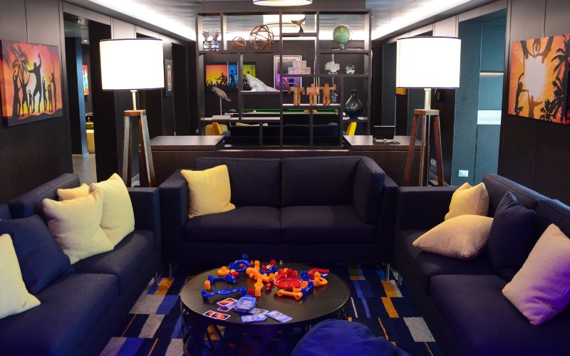 Teens club room on P&O Cruises, featuring comfortable navy blue sofas with yellow pillows, modern decor, and a variety of board games and activities on a coffee table. The room has a cozy and stylish ambiance, with shelving units and colorful artwork creating a welcoming space for teenagers to relax and socialize.