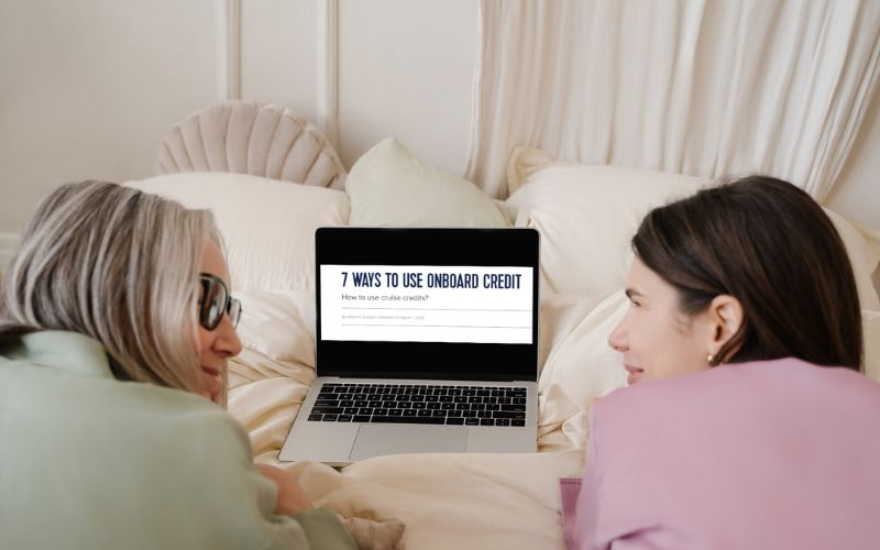 The image shows two women lying on a bed, looking at a laptop screen that displays an article titled "7 Ways to Use Onboard Credit." They appear engaged and interested, suggesting a planning session for a cruise vacation. The setting is cozy and intimate, perfect for a relaxed discussion about maximizing cruise travel benefits.