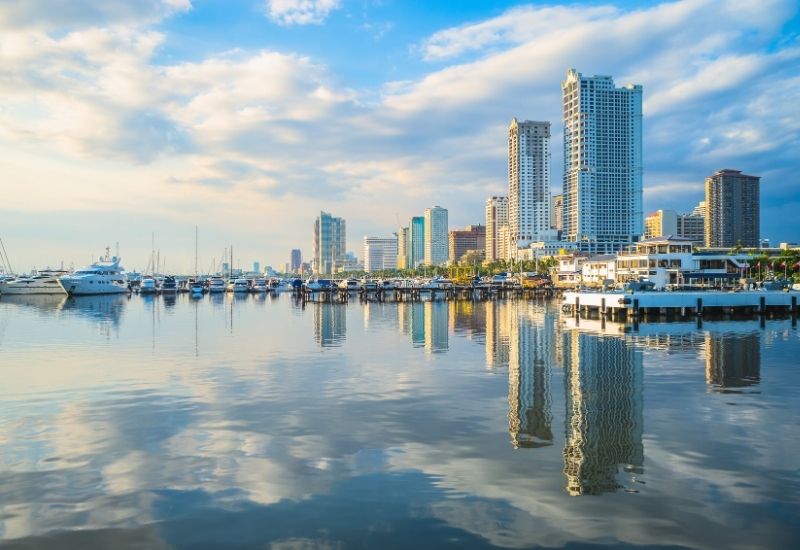 This image depicts the Port of Manila Bay in the Philippines, featuring sleek modern skyscrapers and a marina filled with yachts and boats. The calm waters of the bay beautifully reflect the cityscape and cloudy blue sky, creating a serene and urban coastal ambiance. The view highlights the vibrancy and sophistication of this bustling waterfront hub.