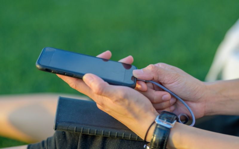 A person is holding a smartphone connected to a portable charger by a cable. The hands are resting on a book with a grassy background, suggesting an outdoor setting. The individual is wearing a black wristwatch, indicating they are using the portable charger to keep their device powered while away from a traditional power source.