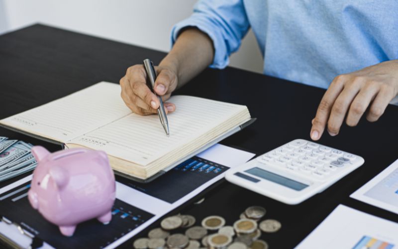 A person is seated at a desk, working on financial planning. They are writing in a notebook with a pen while using a calculator. The desk is scattered with coins, a pink piggy bank, dollar bills, and financial documents, all suggesting a focus on saving money and budgeting. The scene reflects a careful and organized approach to managing personal finances.