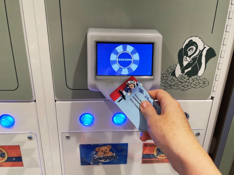 A modern self-service laundry facility on a Disney cruise ship, focusing on a user interaction with the system. A hand is shown using a Disney cruise room card to activate one of the laundry machines. The card reader displays a "CHECKING" message on its screen, indicating the process of validating access. Below the card reader, a variety of laundry detergent and fabric softener options, including "Snuggle," are available in a vending setup, highlighting the convenience offered to guests. The area is equipped with LED lights and digital interfaces, ensuring a user-friendly and efficient laundry experience during the cruise.