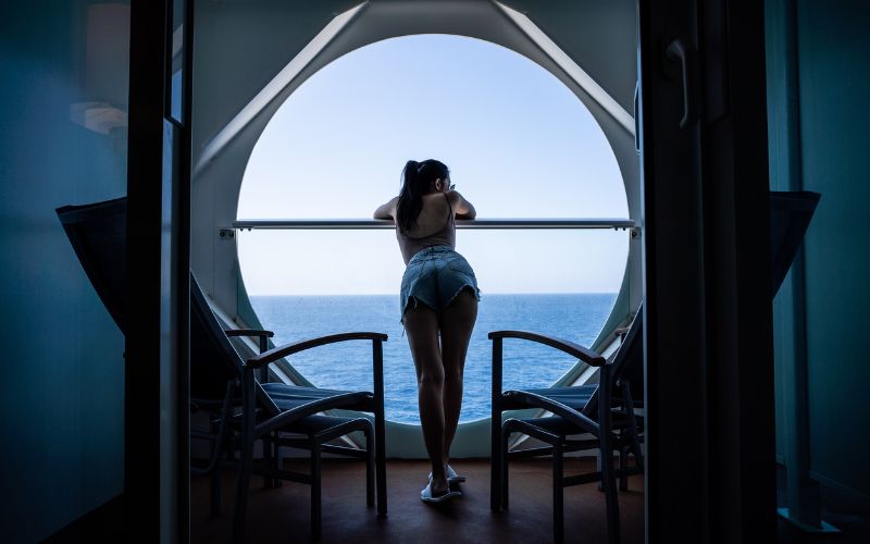 A woman in denim shorts and a tank top is standing on a cruise ship balcony, leaning on the railing as she looks out at the vast ocean through a large, circular window. Two empty chairs are placed beside her, framing the serene view of the open sea under a clear sky.