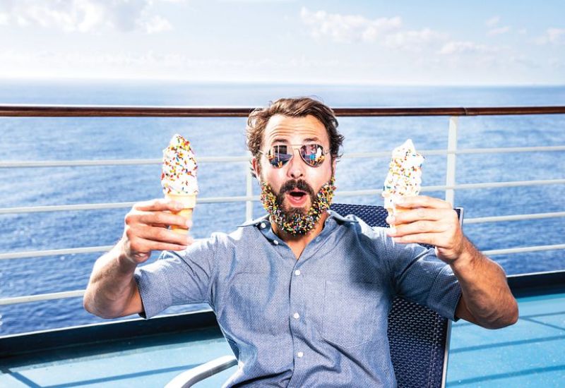 A man with a colourful beaded beard, expressing surprise and delight, holding two ice cream cones on the deck of a cruise ship with the ocean in the background.