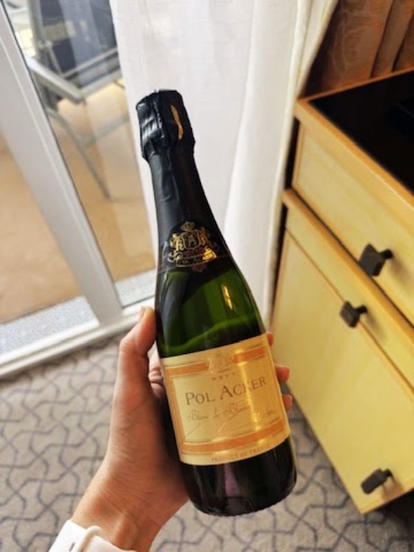 My hand holds a bottle of Pol Acker sparkling wine inside a cabin on the Cunard Queen Mary cruise ship. The background shows part of the cabin with light wooden furniture and a balcony with a glass door.