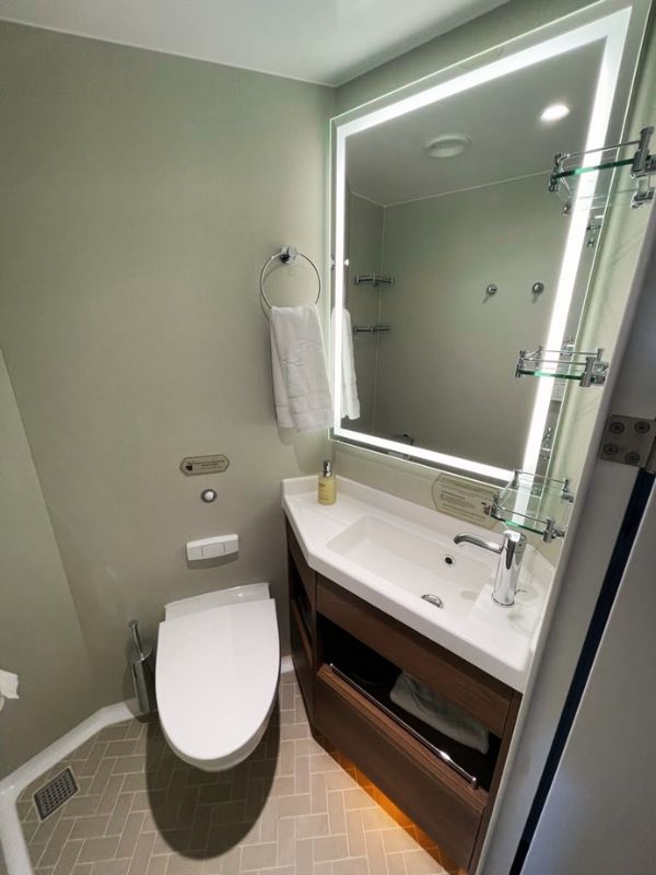 Another section of a split bathroom on a Disney cruise ship, focusing on the toilet area. The design includes a modern, oval-shaped toilet and a rectangular sink housed in a dark wood vanity with storage underneath. Above the sink is a large mirror with integrated lighting, which adds a bright and spacious feel to the compact area. The walls are painted in a soft gray tone, and the floor is tiled in a gray herringbone pattern, maintaining a consistent style with the previous bathroom section.
