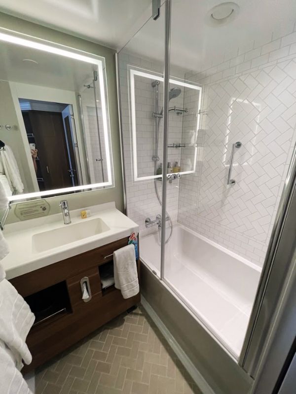 A neatly arranged bathroom with modern fixtures on a Disney cruise ship. The space is split into two sections, with a rectangular sink and vanity on the left, featuring a large mirror with integrated lighting. On the right, there's a full-length shower enclosed by glass doors, complemented by white subway tiles and multiple built-in shelves for toiletries. The floor throughout is laid with herringbone-patterned tiles, enhancing the bathroom's contemporary aesthetic.