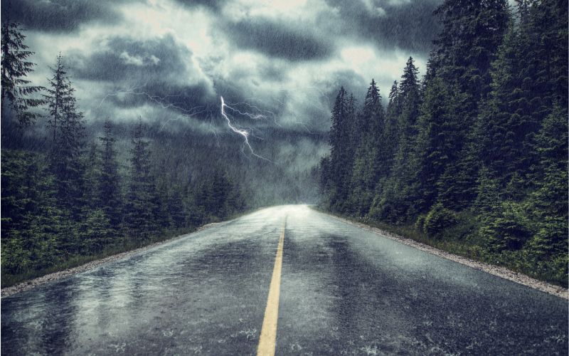 A dramatic scene of a stormy weather with heavy rain and lightning striking over a deserted road, flanked by dense pine forests, emphasizing the intensity and danger of driving in such severe conditions.