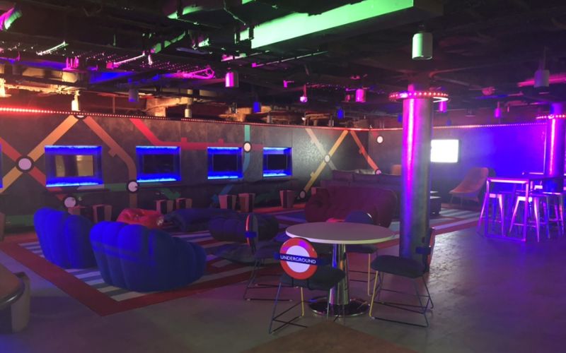 A trendy teen lounge featuring neon lighting, vibrant seating options, and modern decor reminiscent of an urban underground scene. The walls are decorated with a subway map theme, complete with illuminated displays and cushy lounge areas. High tables and chairs provide additional seating, making it a perfect spot for socializing and relaxation.