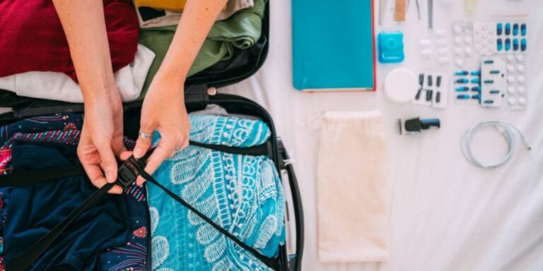A person is seen packing a suitcase, securing the contents with a strap. The suitcase contains neatly folded clothing, including a turquoise patterned garment. Next to the suitcase on the bed, various travel essentials are laid out, including medication, toiletries, a book, a phone charger, and other small items. The image conveys preparation and organization for a cruise or trip.