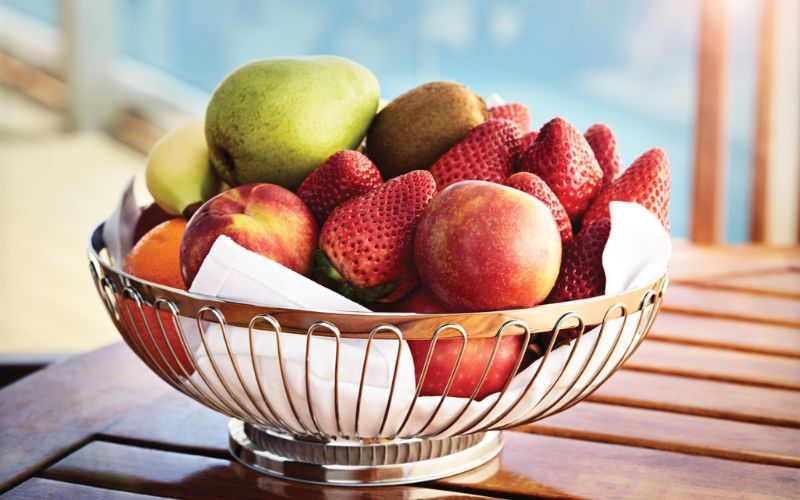 A metal bowl filled with a variety of fresh fruits, including apples, pears, strawberries, and kiwis, is placed on a wooden table on a cruise ship. The background shows a blurred view of the ocean, suggesting a relaxing outdoor dining area.