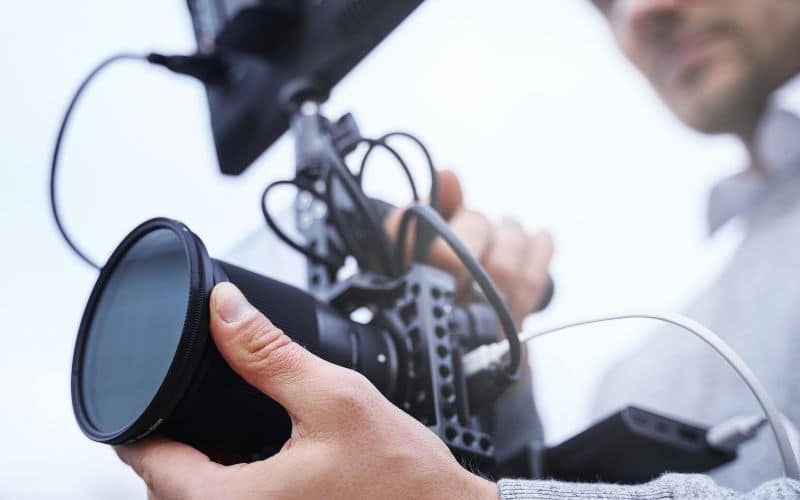A videographer is adjusting the settings on a professional video camera, ensuring the lens and equipment are ready for filming. The image focuses on the camera and the videographer's hands, with the videographer slightly out of focus in the background.