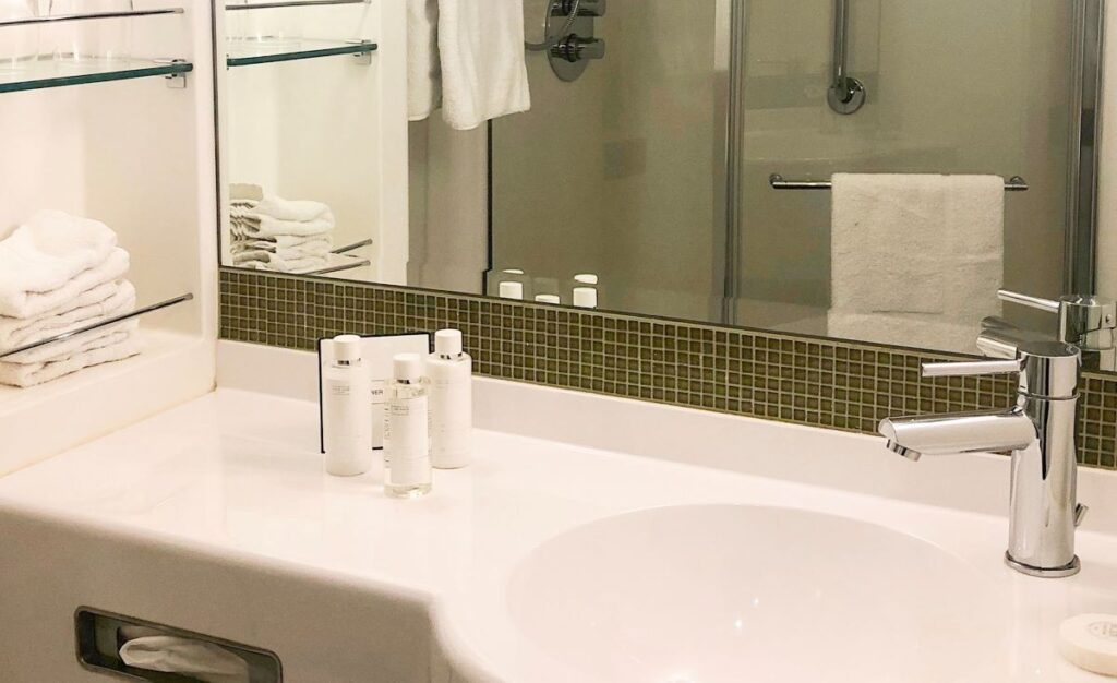 Neatly arranged bathroom featuring a sleek white sink and chrome faucet, with White Company toiletries on the countertop. Glass shelves with stacked towels and a mirror reflect the shower area in the background on a P&O cruise ship.