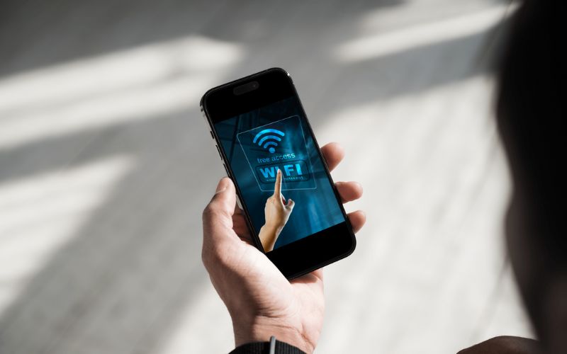 The image shows a close-up of a person holding a smartphone displaying a screen with a "Free WiFi Access" graphic. A hand on the screen is pointing to the WiFi icon, symbolizing easy connectivity. The photo captures the modern reliance on digital connectivity, especially in mobile settings, and emphasizes the convenience of accessing free WiFi.