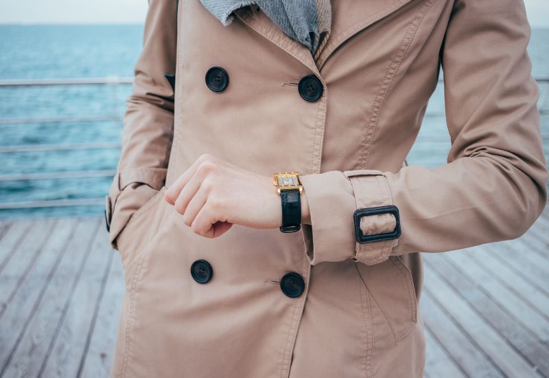 A woman in a beige trench coat and gray scarf stands on a wooden deck near the ocean, checking the time on her wristwatch. Her left hand is in her pocket while her right hand is adjusting the watch, with the sea visible in the background.
