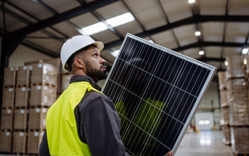 A worker wearing a white helmet and high-visibility jacket carries a large solar panel inside a warehouse. The focus is on his profile as he carefully handles the panel, indicative of his professionalism and the importance of safety in his work environment.
