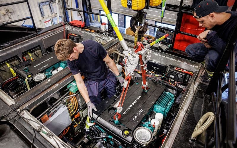 Two technicians work diligently on the engine of a cruise ship, located in a well-equipped workshop. The image captures one worker adjusting components inside the open engine bay, while the other observes carefully, both surrounded by various tools and mechanical parts, highlighting their expertise in marine engineering.