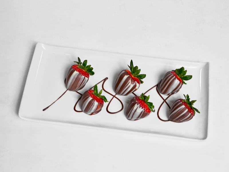 Five fresh strawberries dipped in dark chocolate and drizzled with white chocolate, elegantly presented on a rectangular white plate against a minimalist background.
